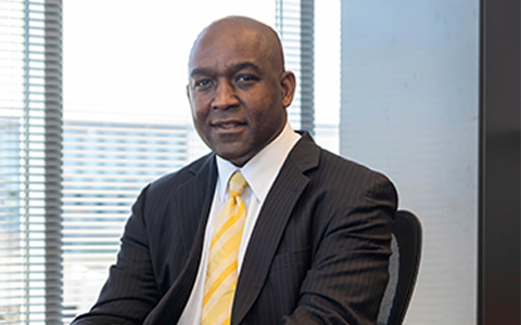 Reggie Scales sits at his desk in a black sports coat and yellow tie.