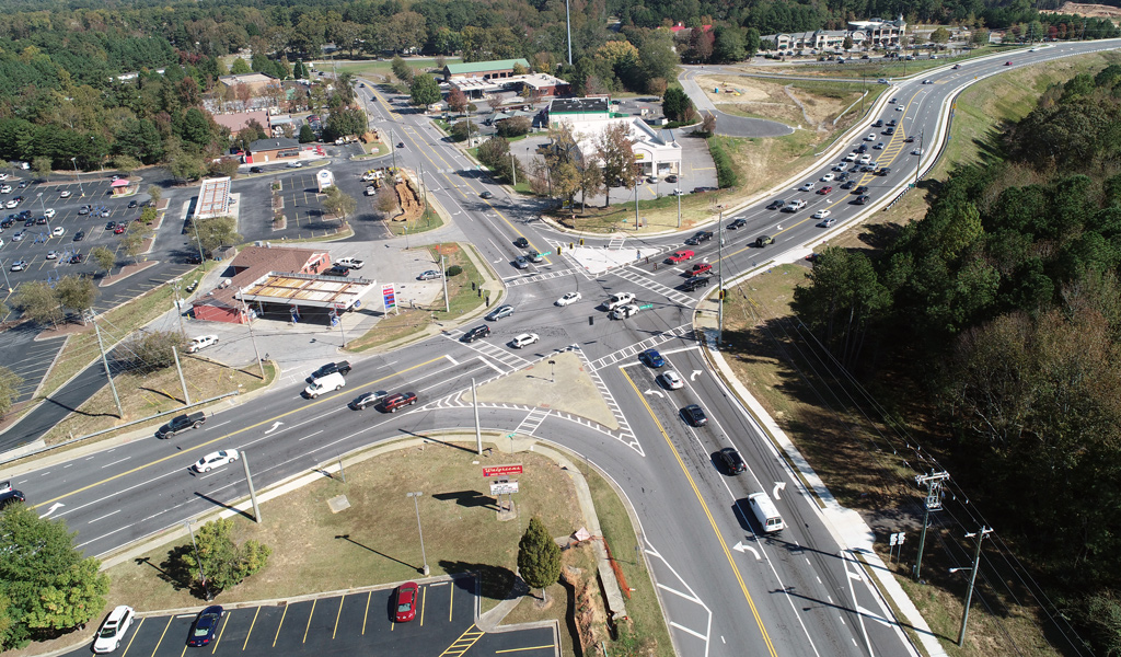 An aerial photograph depicts a multi-lane roadway interchange with vehicles traveling on it and trees and buildings located beside the roadway.