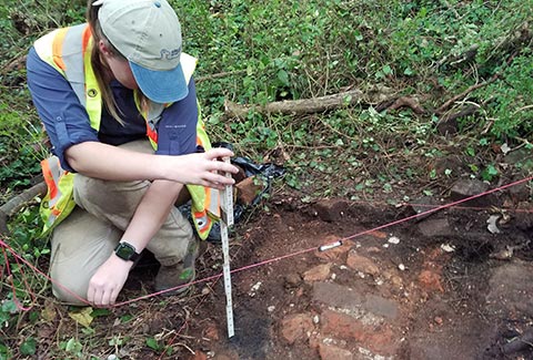 A woman kneels near a hole in the ground and measures the depth.