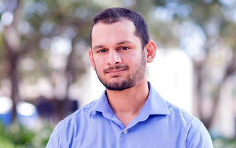 A business portrait of a smiling man with dark hair and a blue button-down shirt in an outdoor setting.