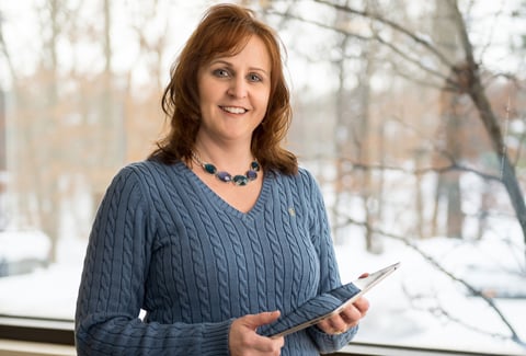 Wendy wearing a blue sweater and holding an iPad while standing in front of a window.