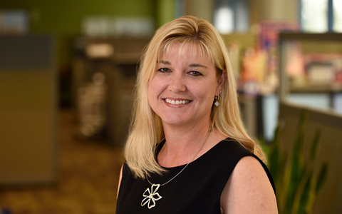 A business portrait of a smiling woman with blonde hair wearing a flower necklace in an office setting.