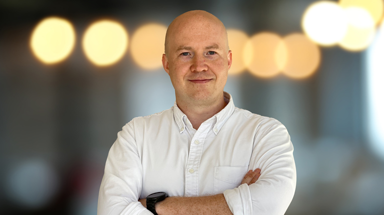 Andrew wearing a white shirt standing with his arms folded. 