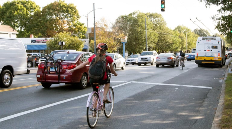 A woman wearing a bicycle helmet and riding a bike in a bike lane uses hand signals on a busy road.