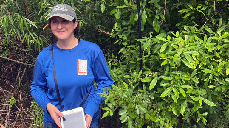 A woman in a long sleeve blue shirt, jeans, and ball cap holds an iPad and stands beside a pole used to collect acoustic data from bats