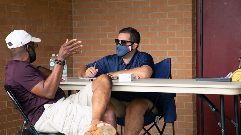 Two men wearing masks sit at a folding table outside a brick building while one man takes notes