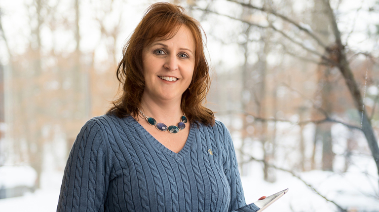 Wendy wearing a blue sweater and holding an iPad while standing in front of a window.