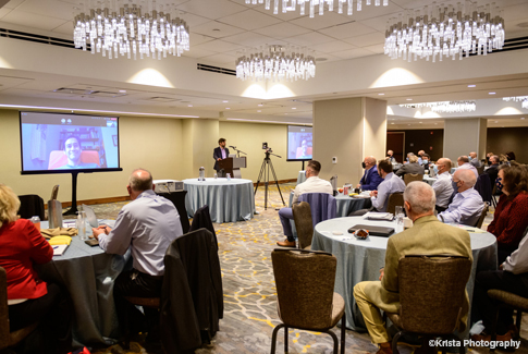 A group of people listen to speakers at a podium and via remote access.