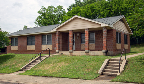 A red brick one story residence is pictured with two sets of concrete stairs leading up to the yard and entrance. 