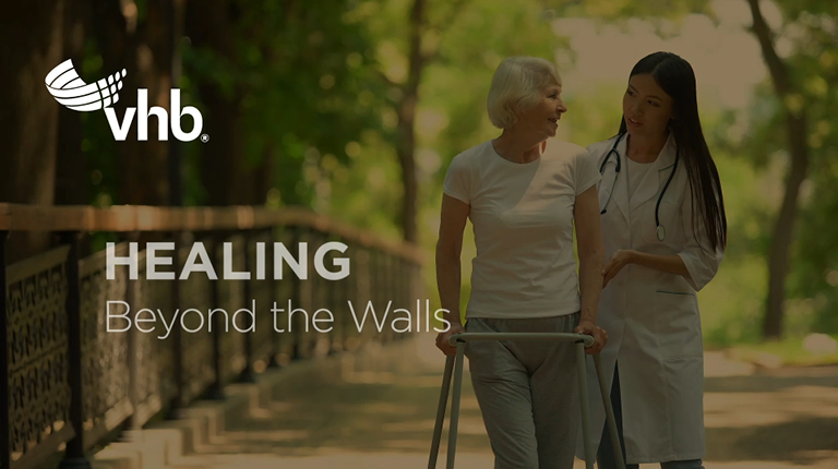 A woman wearing a medical professional lab coat is helping an elderly woman who is using a walker to talk a walk outdoors